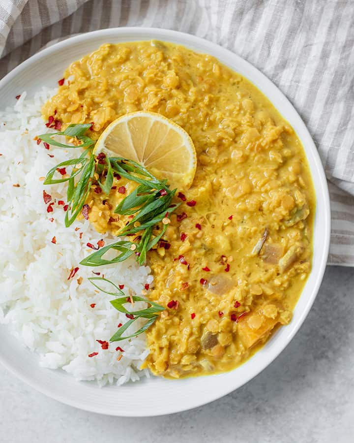 Plate of white Jasmine rice with curry served on the side garnished with scallions, red pepper flakes and a wedge of lemon.