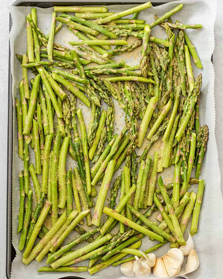 Asparagus seasoned and ready for baking along with some garlic cloves on a baking tray.
