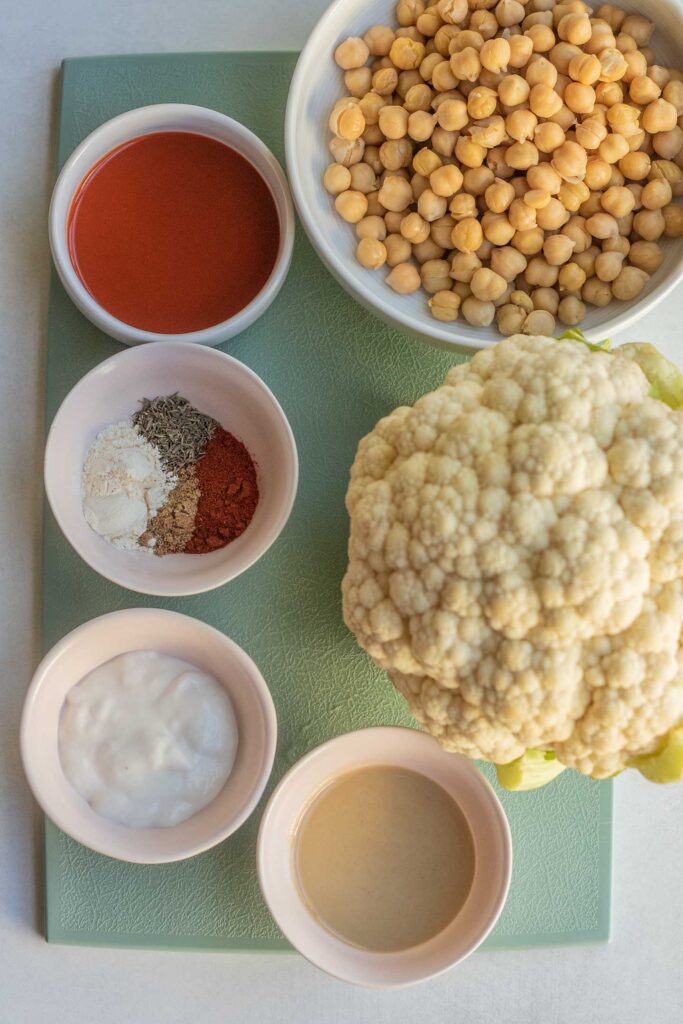 Blue cutting board topped with a head of cauliflower, bowl of chickpeas, buffalo sauce, spices, yogurt and tahini.