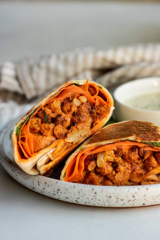 Halved buffalo chickpea burrito, stacked on eachother on a white plate with bowl of dip on the side.