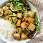 Plate of tofu, white rice with greens and zucchini set on a striped napkin.