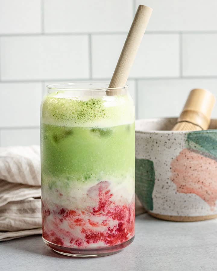 Glass of matcha, milk and strawberries with a straw and matcha bowl in the background.