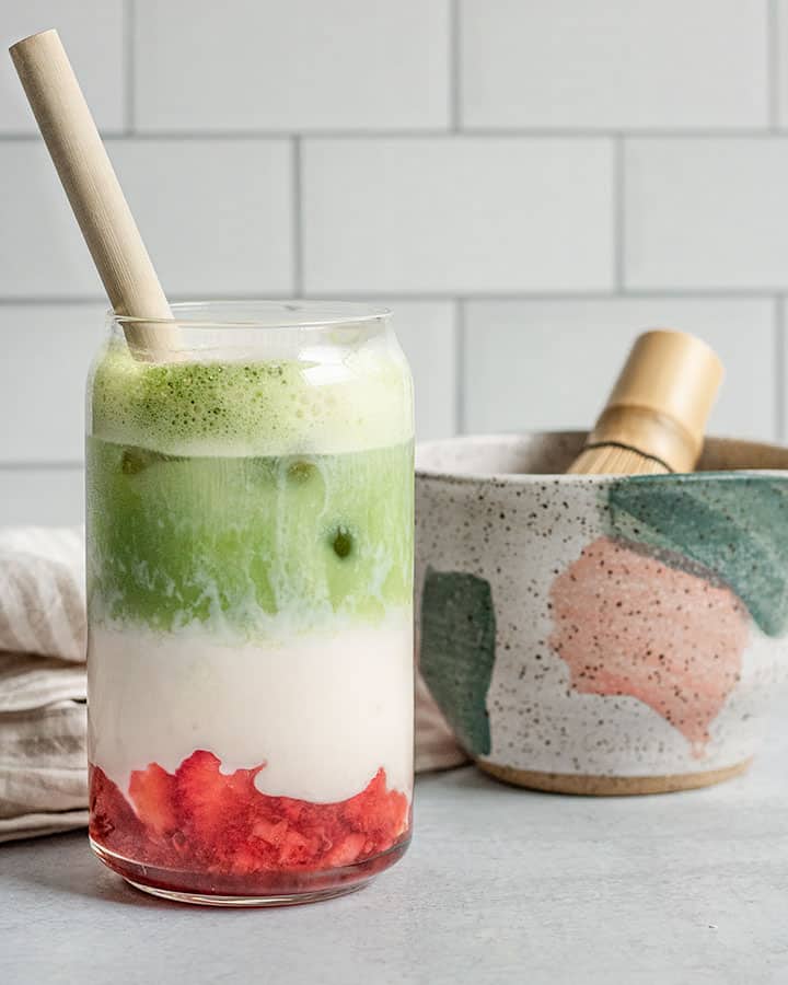 Separated strawberries, milk, and matcha topped with milk foam in a tall glass with a straw.