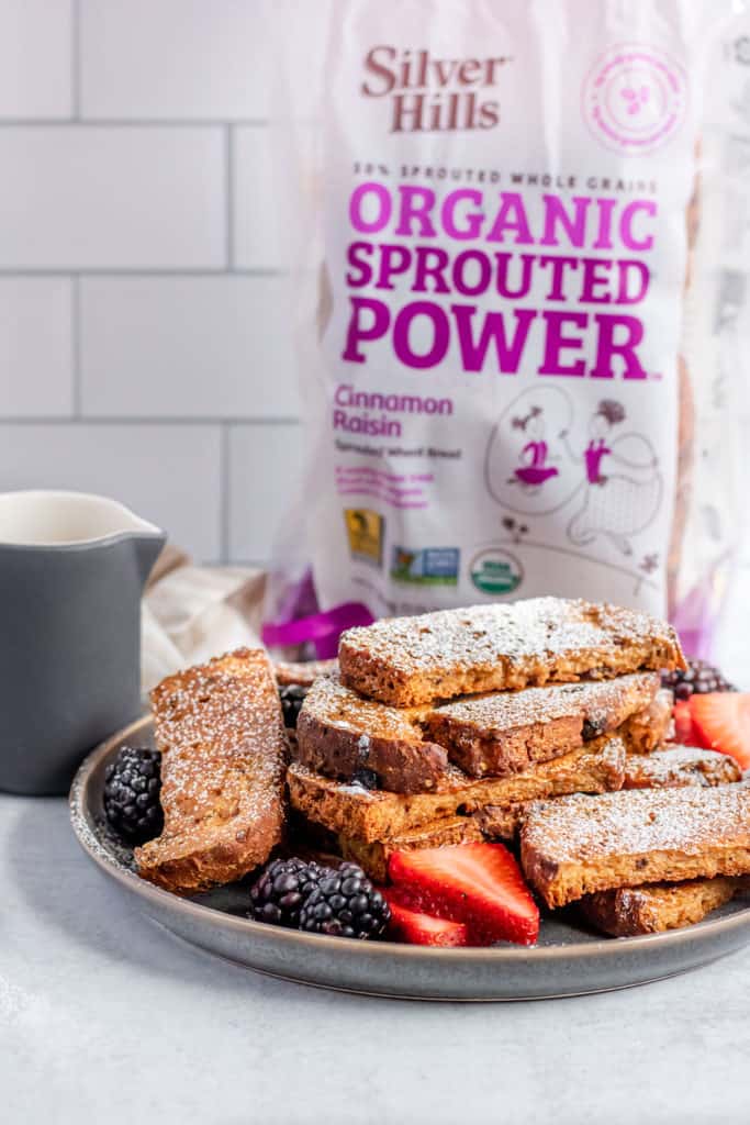 French toast sticks on a plate dusted with powdered sugar and a bag of cinnamon raisin bread in the back.