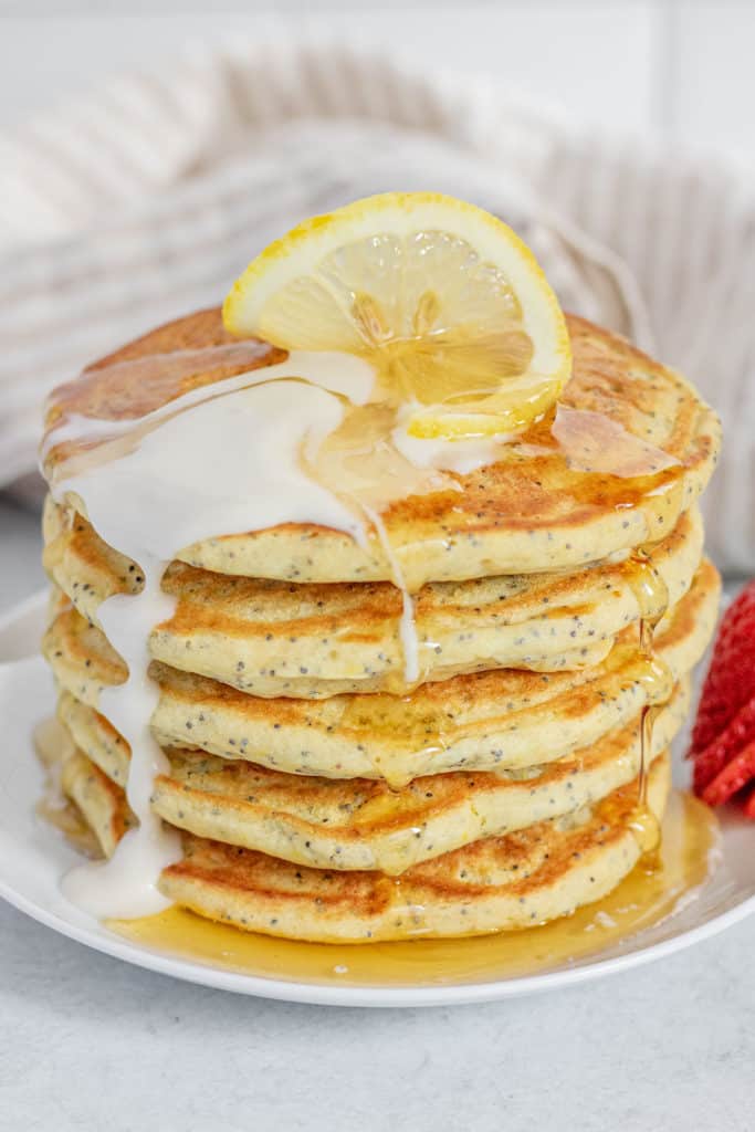 Stack of lemon poppy seed pancakes topped with a slice of lemon, vegan yogurt and maple syrup.
