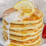 Pouring syrup over a stack of pancakes topped with yogurt and lemon slice.