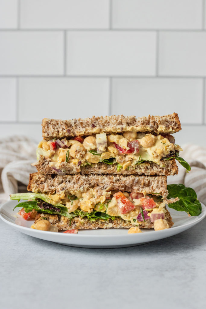 Chickpea salad sandwich cut in half ono a white plate in front of tiled background.