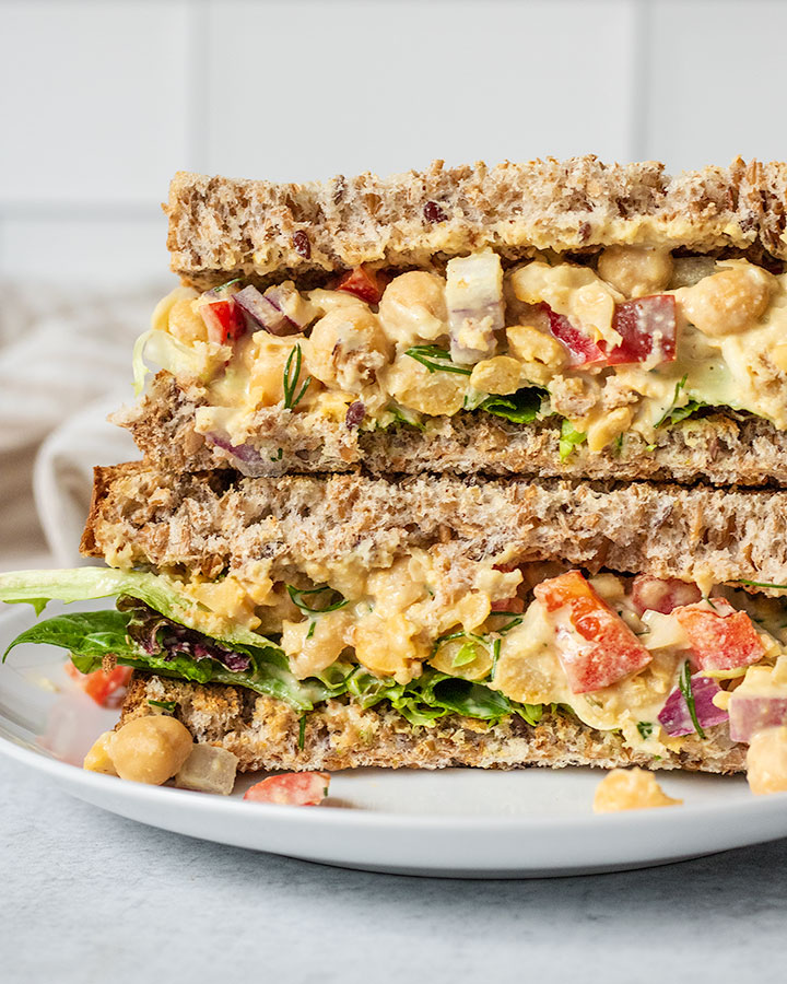 Chickpea salad sandwich fully assembled on whole grain bread and added greens on a plate.