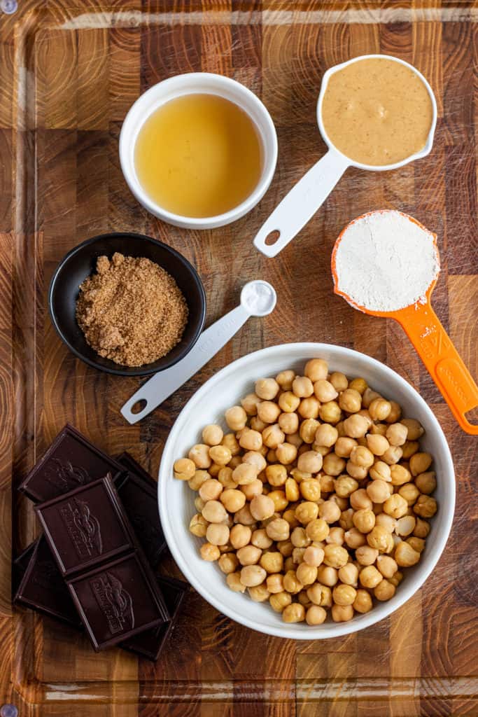 Ingredients for cookie dough bites set on a cuttin board including chickpeas, oat flour, peanut butter, maple syrup, brown sugar and dark chocolate.