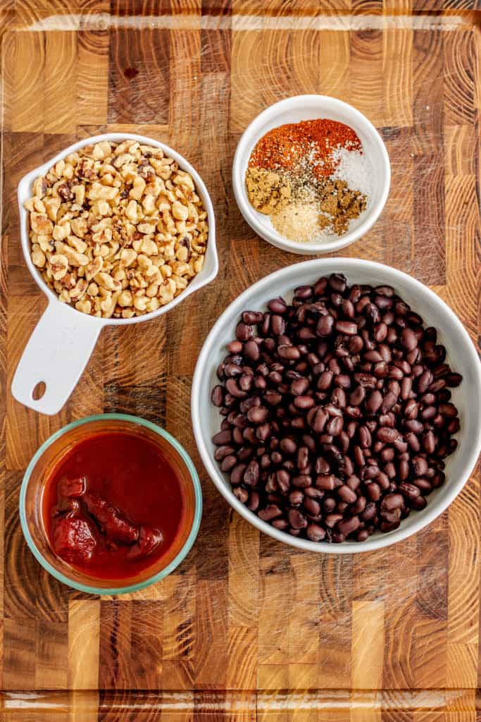 Cutting board with chipotle peppers, walnuts, black beans and spices in separate bowls.