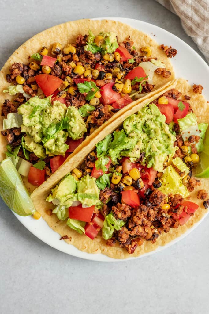 Plate with 2 tacos layered with lettuce, vegan taco meat, tomatoes, corn and mashed avocado.
