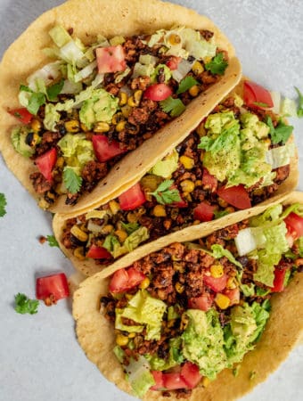 Top down image of three tacos placed next to each other and topped with avocado, tomato and cilantro.