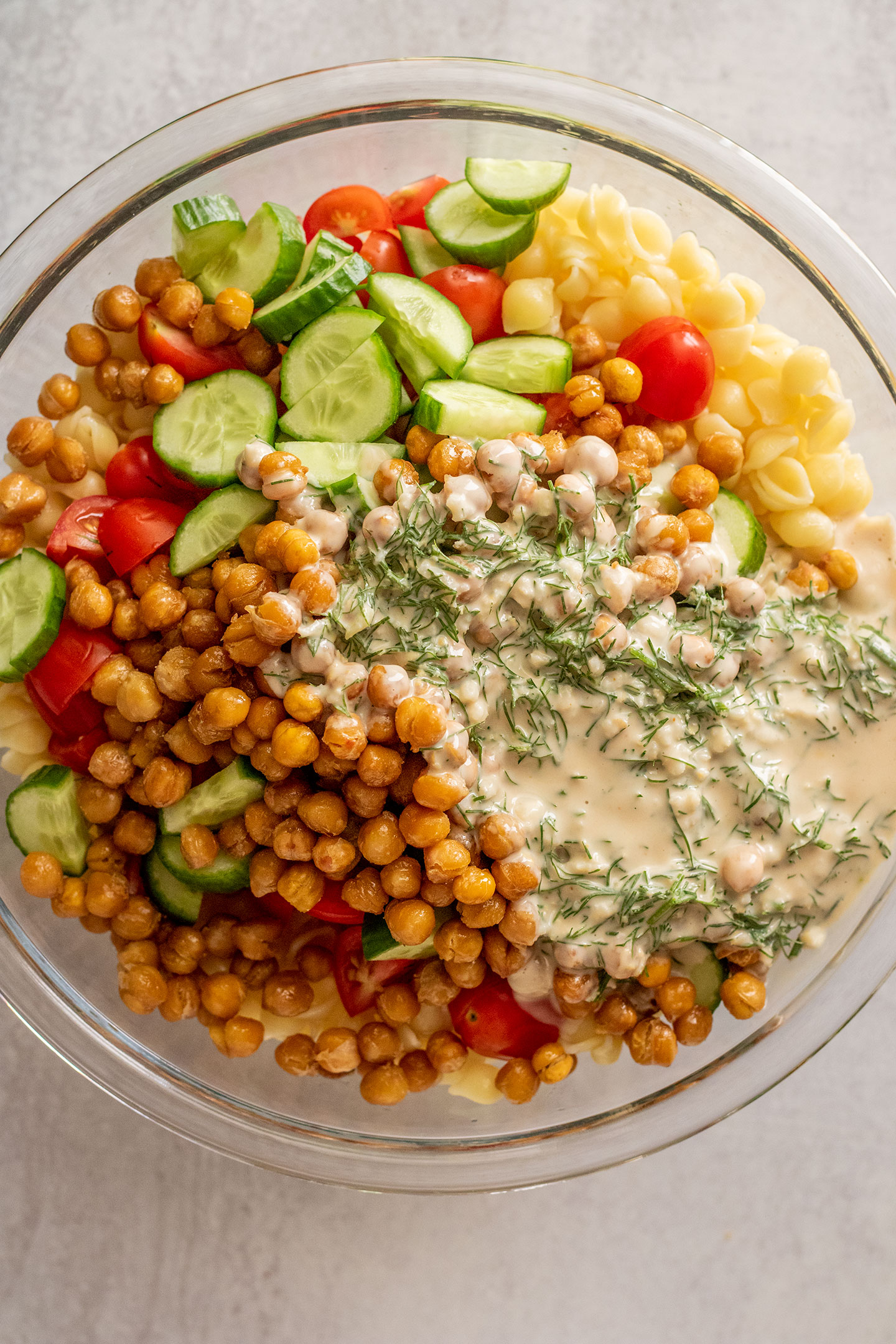 Combining the pasta, tomatoes, cucumbers and roasted chickpeas with the creamy dressing in a bowl.