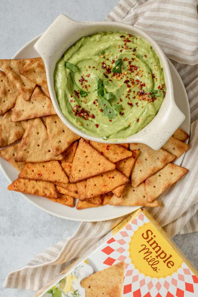 Veggie Pita Crackers spread around a plate next to a bowl of avocado dip.