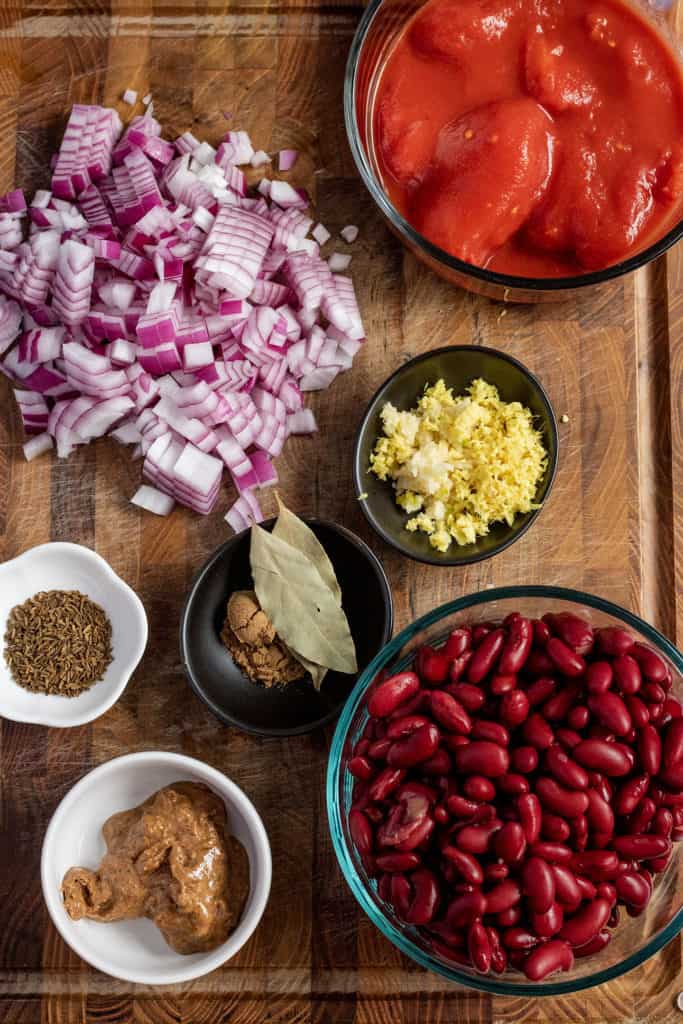 Cutting board topped with diced onions, cumin seeds, tomatoes, ginger, garlic, spices and red beans.