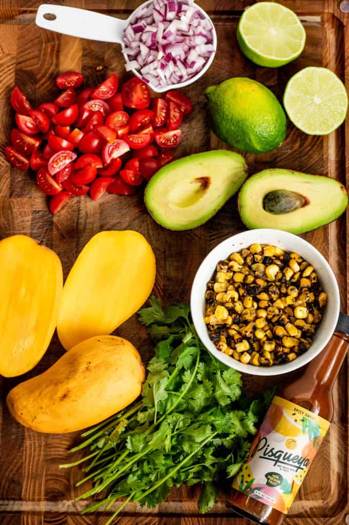 Mango, avocado, cherry tomatoes, lime, cilantro, corn, onion and hot sauce placed together on a cutting board.
