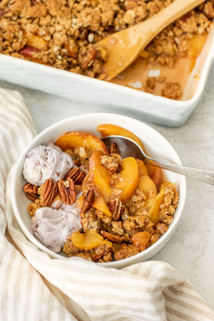 Side view of bowl of peach crisp next to tray of baked peach crisp.