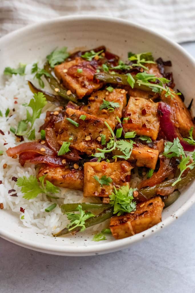 Side view of a bowl of sweet and spicy tempeh topped with cilantro and chives over a bed of rice.