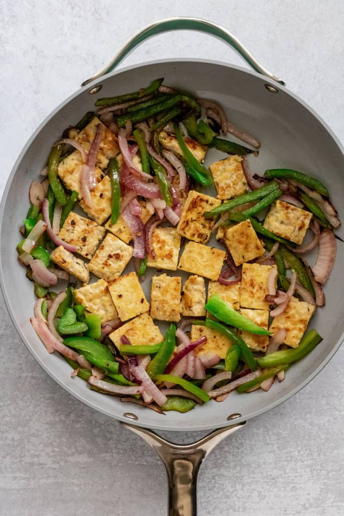 Tempeh browning next to peppers and onions in a large skillet pan.