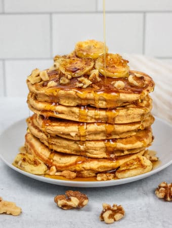 Close up view of banana nut pancakes topped with walnuts and caramelized banana being drizzled with maple syrup.