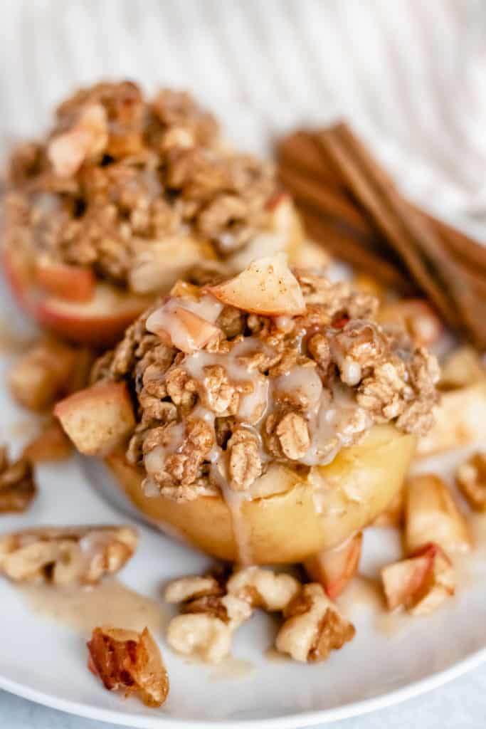 Two baked apples with oat crumble on a plate with cinnamon sticks in the background.