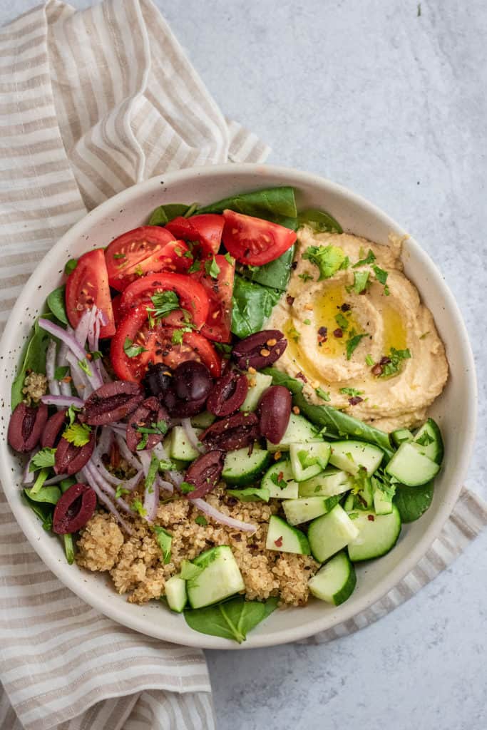 Fully assembled nourish bowl with hummus, cucumbers, tomatoes, onions, olives, and quinoa.