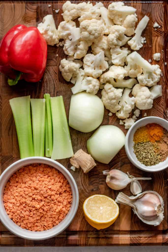 Cutting board with red lentils, bell pepper, onion, cauliflower florets, celery, ginger, garlic cloves, spices and half a lemon.