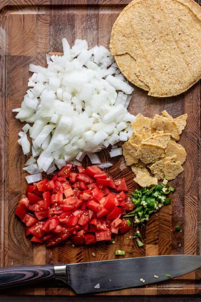 Cutting board with chopped onions, tomatoes and serrano peppers along with some torn tortillas.