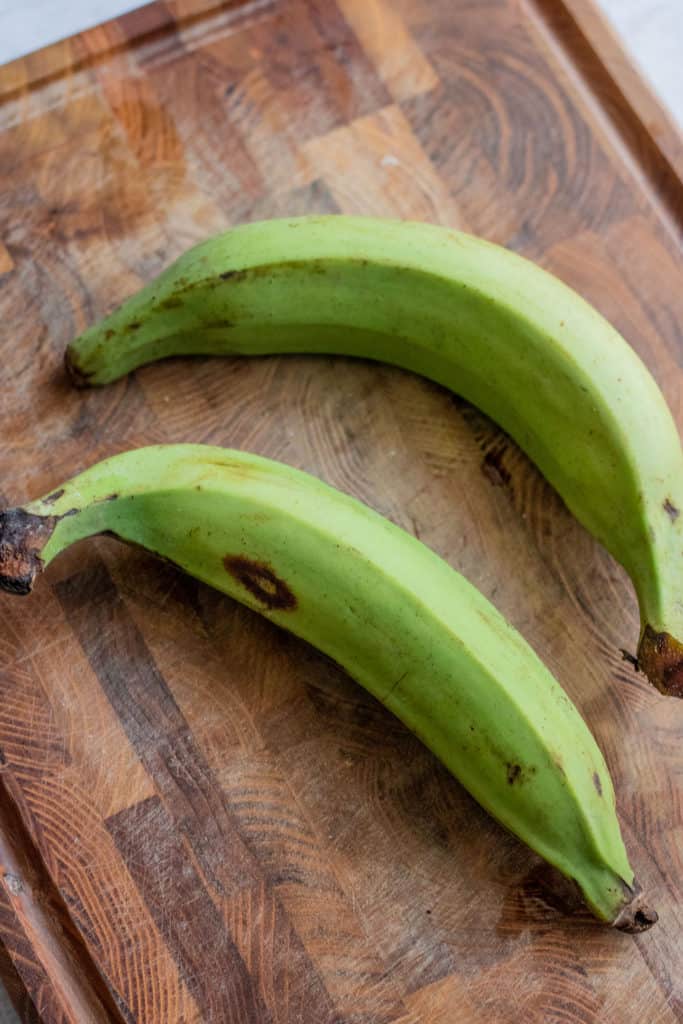 Two whole green plantains laid out on a wooden cutting board.