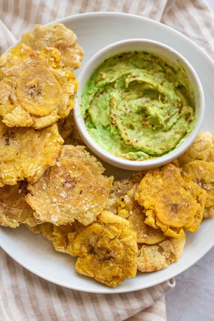 Top down shot of plantains on a white plate next to some mashed avocado.