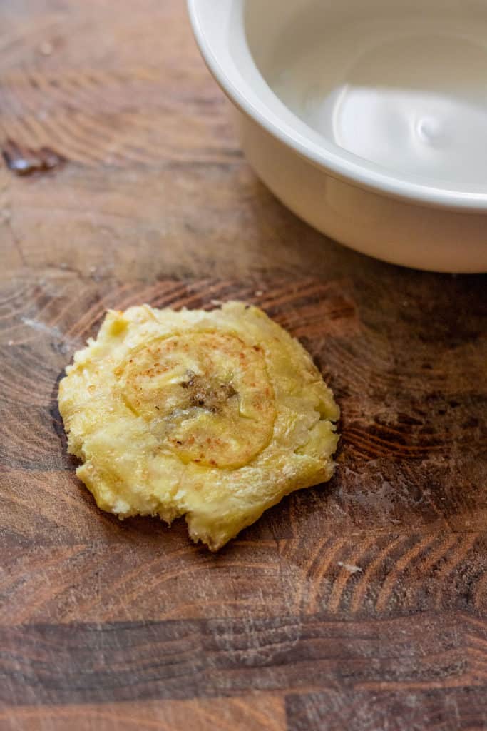 Smashing the warm plantain with a flat bottomed dish on a cutting board.