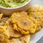 A plate of air fried plantains on a white plate set next to some mashed avocado in a white bowl.