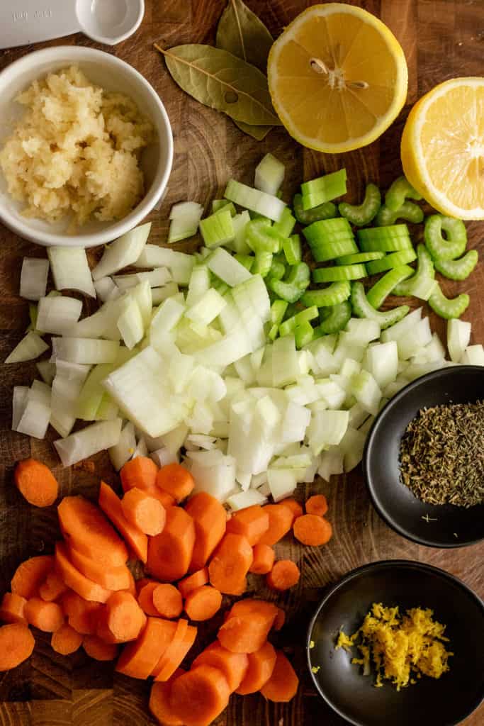 Cutting board with chopped onions, garlic, carrots and celery with herbs and lemon in small dishes.