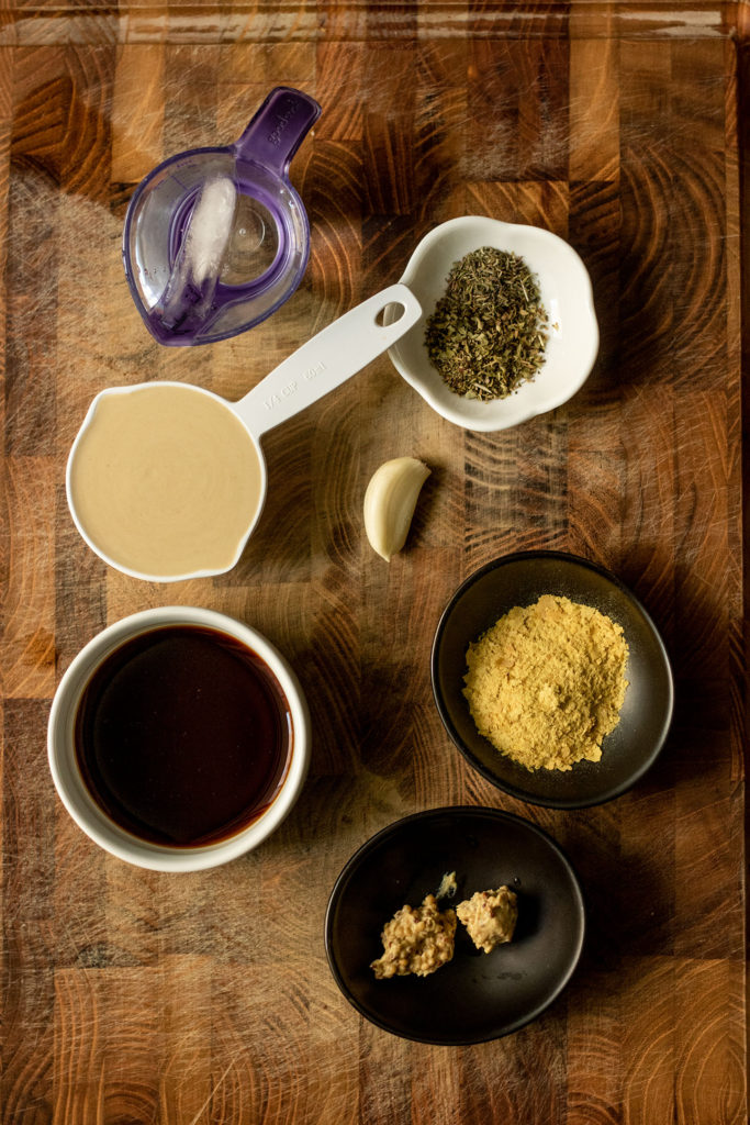 All the dressing ingredients placed on a wooden cutting board including herbs, tahini, balsamic vinegar, nutritional yeast and mustard.
