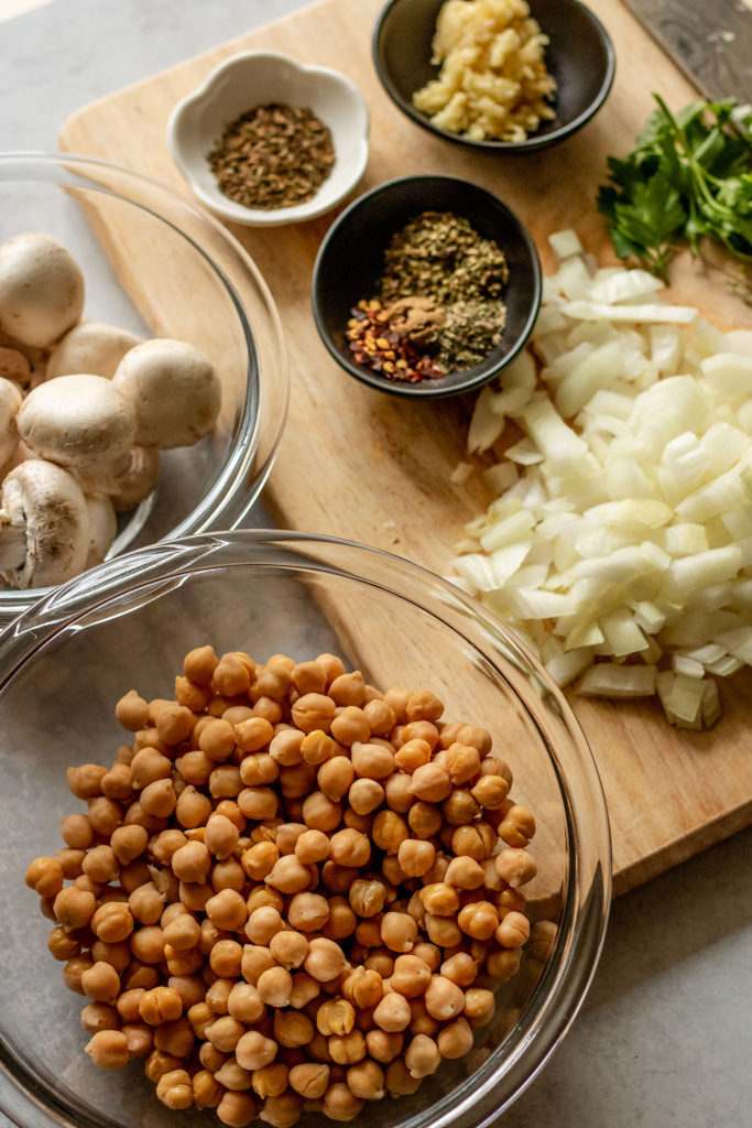 Cutting board with diced onions, spices, herbs and garlic along with a bowl of mushrooms and chickpeas.