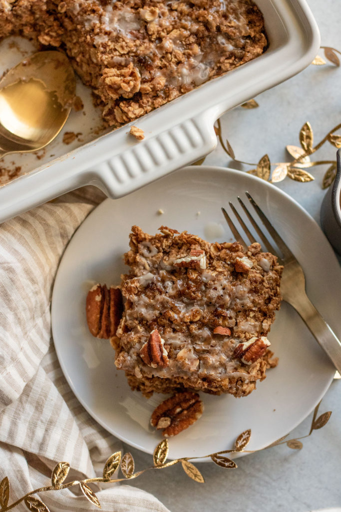 Serving a square of baked oatmeal on a plate with icing and pecans on top.