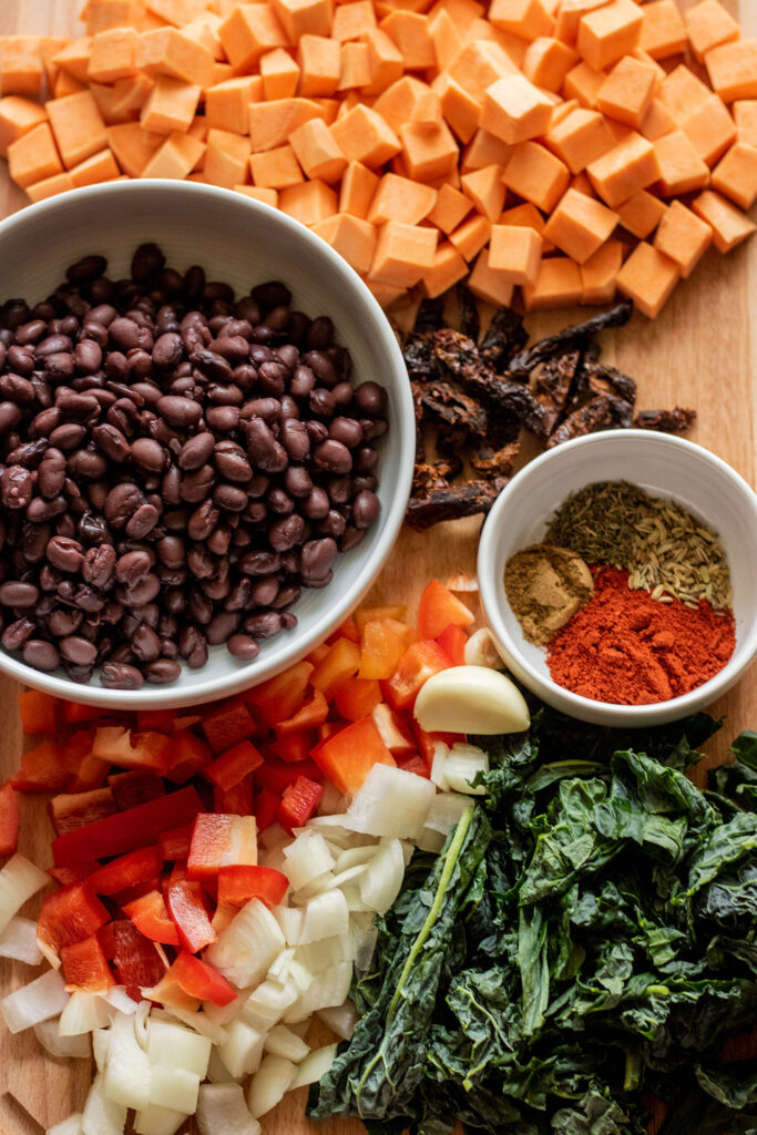 Sweet potatoes, black beans, sun-dried tomatoes, seasonings, garlic and veggies on a cutting board.