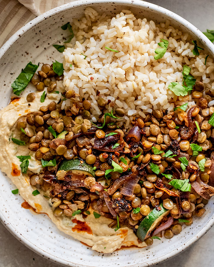 Close up of a bowl of lentils on top of hummus and served with rice and a drizzle of chili oil.