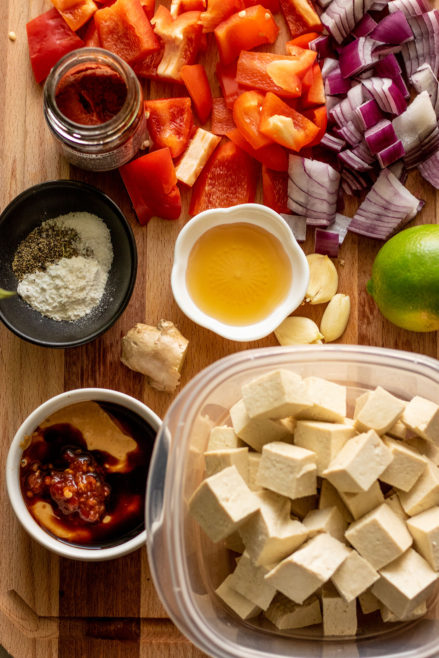 All the curry ingredients on a cutting board including red curry paste, pepper, onion, peanut butter, soy sauce, tofu, lime, ginger and garlic.