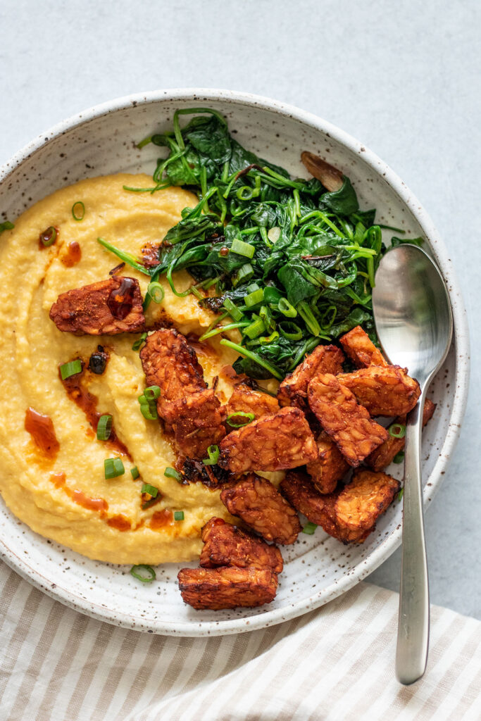 Top view of a bowl of polenta topped with spinach, tempeh and green onions.