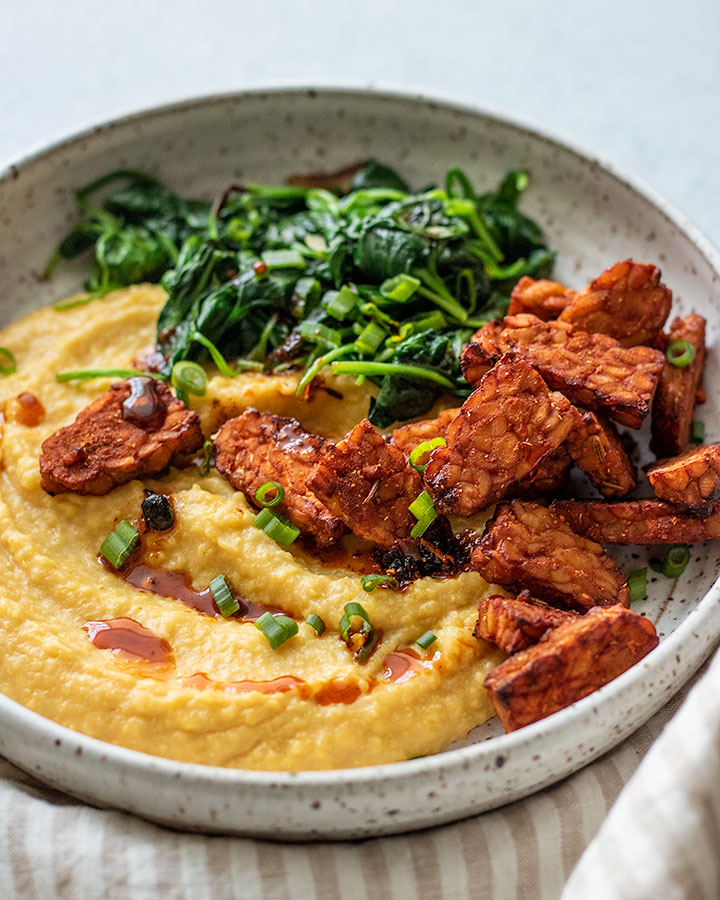 Side view of a bowl of polenta topped with air fried tempeh and sautéed spinach.