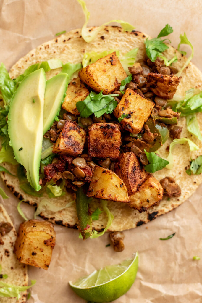 A single taco on parchment paper topped with lentils, roasted potatoes and avocado slices.