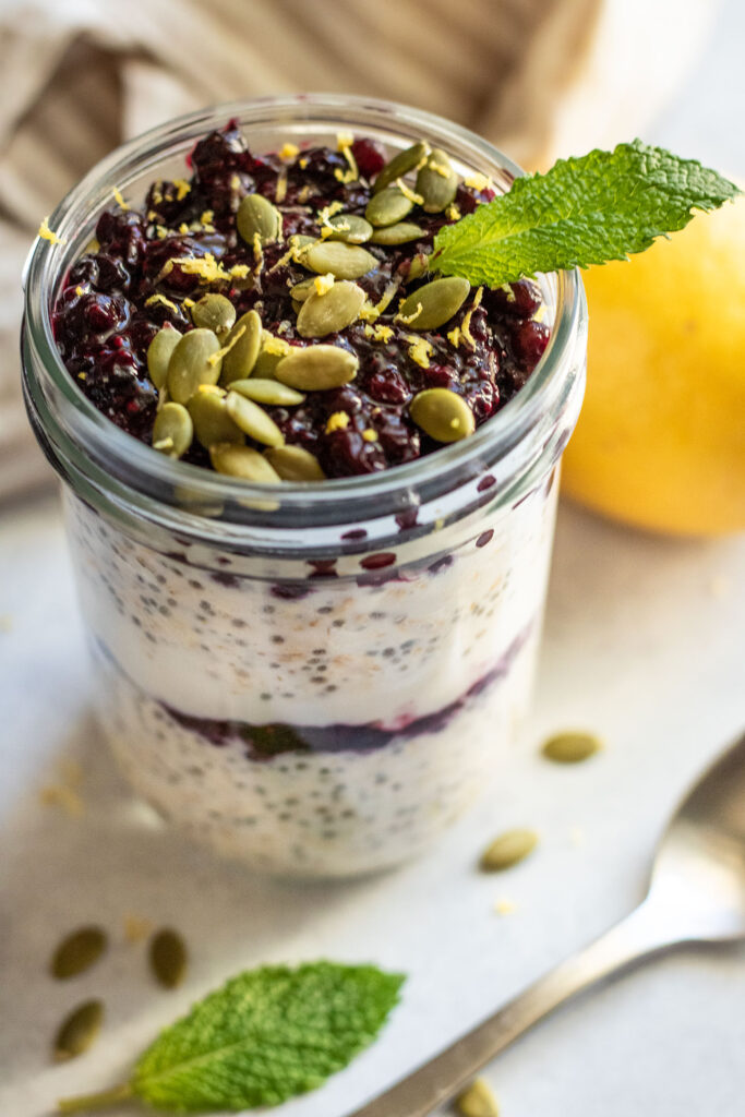Side top view of a jar of overnight oats topped with blueberry jam, lemon zest, pepitas and mint.