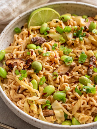 Close up of a bowl of noodles tossed with peanut sauce, edamame, cabbage and mushrooms then served with a lime.