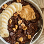 Close up of a bowl of fudgy chocolate oatmeal topped with banana, nut butter, chocolate chips and hazelnuts.