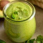 Close up view of a jar of cilantro sauce topped with cilantro leaves sitting next to an avocado and jalapeno.
