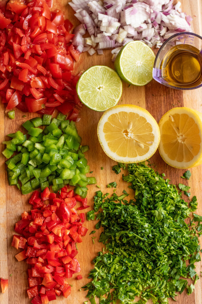 Finely diced tomato, peppers, parsley, and sliced lemon and lime on a cutting board.