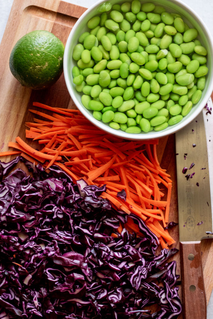 Cutting board with edamame, lime, carrots and sliced cabbage.