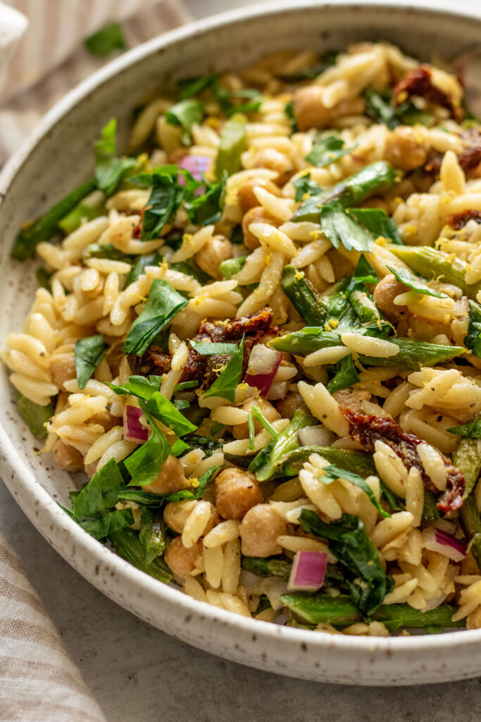 Side view of a bowl of orzo mixed with herbs, sun-dried tomatoes, asparagus and chickpeas.