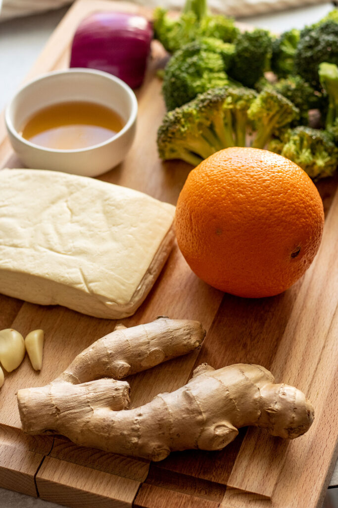 Cutting board with an orange, broccoli florets, tofu, ginger, garlic and maple syrup on it.